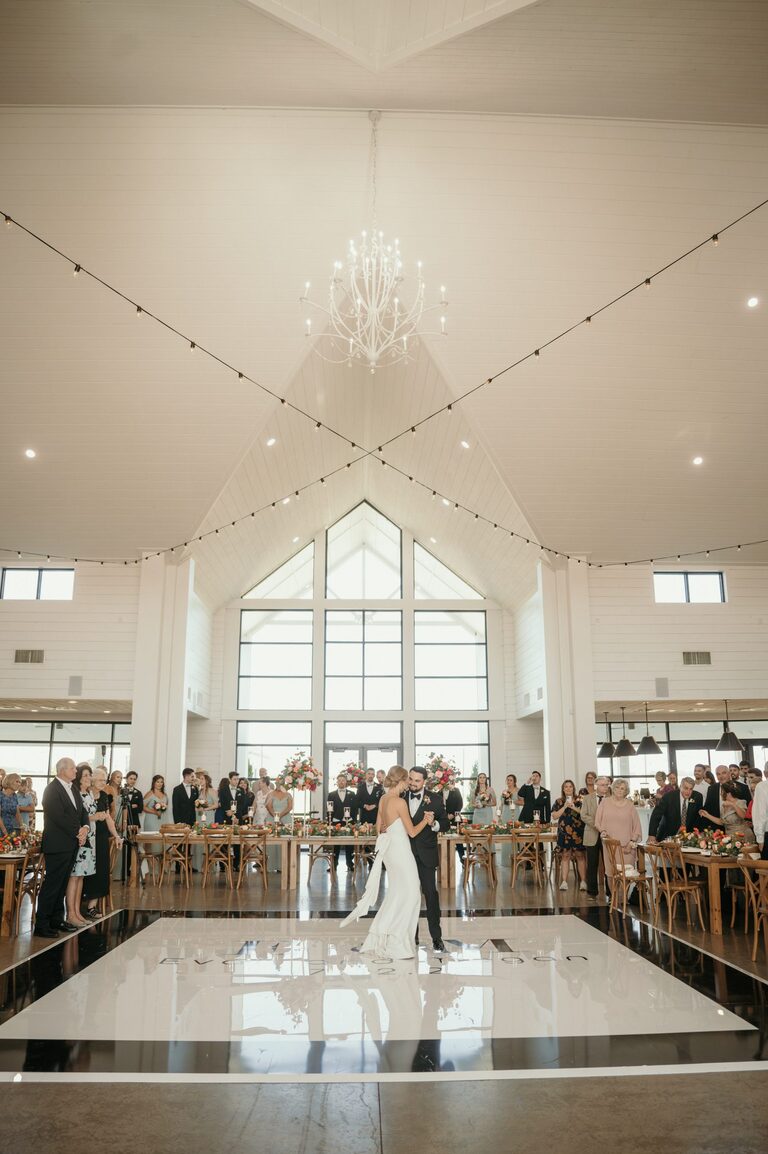 Bride and groom dancing beneath a chandelier as guests watch from around the dance floor at The Gardenia Venue.